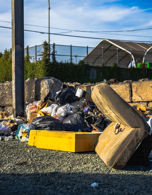 A close-up view of an unmanaged rubbish pile situated on a gravel surface, consisting of various waste materials including black plastic rubbish bags, a broken and stained beige car tyre, and a yellow plastic container. The waste appears to include household and discarded items, with some bags partially torn open revealing contents such as paper and packaging. Behind the pile is a low stone wall made of large, roughly cut stones, with greenery and trees visible above it. In the background, a wire fence and a shed or covered structure with a curved metal roof can be seen, along with utility poles and electrical wires against a partly cloudy sky. The scene conveys an environment awaiting curbside or independent waste collection, fitting within a context of private rubbish removal services like those offered by Rubbish Removal Tufnell Park in the area.