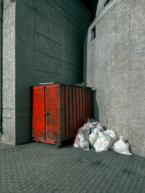 A close-up view of an unmanaged rubbish pile situated on a gravel surface, consisting of various waste materials including black plastic rubbish bags, a broken and stained beige car tyre, and a yellow plastic container. The waste appears to include household and discarded items, with some bags partially torn open revealing contents such as paper and packaging. Behind the pile is a low stone wall made of large, roughly cut stones, with greenery and trees visible above it. In the background, a wire fence and a shed or covered structure with a curved metal roof can be seen, along with utility poles and electrical wires against a partly cloudy sky. The scene conveys an environment awaiting curbside or independent waste collection, fitting within a context of private rubbish removal services like those offered by Rubbish Removal Tufnell Park in the area.