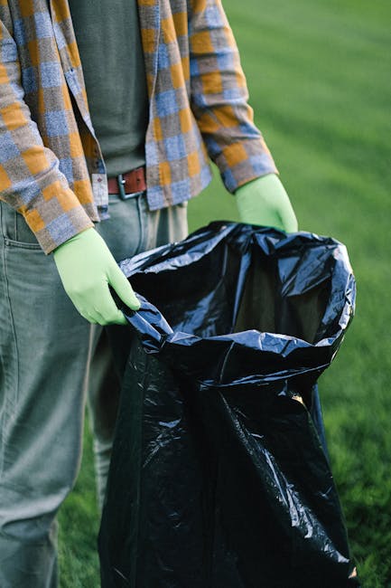 A person standing outdoors on a grassy area is holding open a large black plastic rubbish bag, prepared for waste collection or disposal; they are wearing a green glove on each hand, a casual outfit with a plaid shirt in shades of yellow, grey, and orange over a dark green t-shirt, and grey trousers with a brown belt. The background shows a well-maintained, vibrant green lawn, and the lighting suggests daytime with natural light illuminating the scene. The image depicts a typical scene of rubbish collection, which may relate to private waste handling or alternative waste clearance services offered by companies like Rubbish Removal Tufnell Park, supporting the need for on-site rubbish management without relying solely on local authority collections.