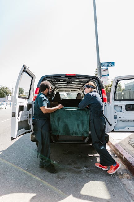 Two waste removal specialists from Rubbish Removal Tufnell Park are loading a large, dark green waste container into the back of a white panel van. The container appears to be made of a heavy-duty, textured material, possibly plastic or composite, with visible ridges along its sides. The van's rear doors are wide open to facilitate loading, revealing a spacious interior. The specialists are wearing dark uniforms; one has a cap and gloves, the other is wearing sunglasses and sneakers. They are positioned on a paved street beside a curb, with a clear, bright sky overhead. In the background, there are street signs, trees, and other vehicles, indicating an urban or suburban environment. The scene captures a tidy, organized waste collection activity, characteristic of private rubbish removal services like Rubbish Removal Tufnell Park, which offers alternative waste handling solutions outside of council collection systems. The overall setting emphasizes on-site clearance and efficient rubbish disposal in a typical residential or mixed-use area.