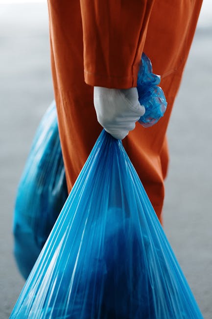 A person standing outdoors on a grassy area is holding open a large black plastic rubbish bag, prepared for waste collection or disposal; they are wearing a green glove on each hand, a casual outfit with a plaid shirt in shades of yellow, grey, and orange over a dark green t-shirt, and grey trousers with a brown belt. The background shows a well-maintained, vibrant green lawn, and the lighting suggests daytime with natural light illuminating the scene. The image depicts a typical scene of rubbish collection, which may relate to private waste handling or alternative waste clearance services offered by companies like Rubbish Removal Tufnell Park, supporting the need for on-site rubbish management without relying solely on local authority collections.